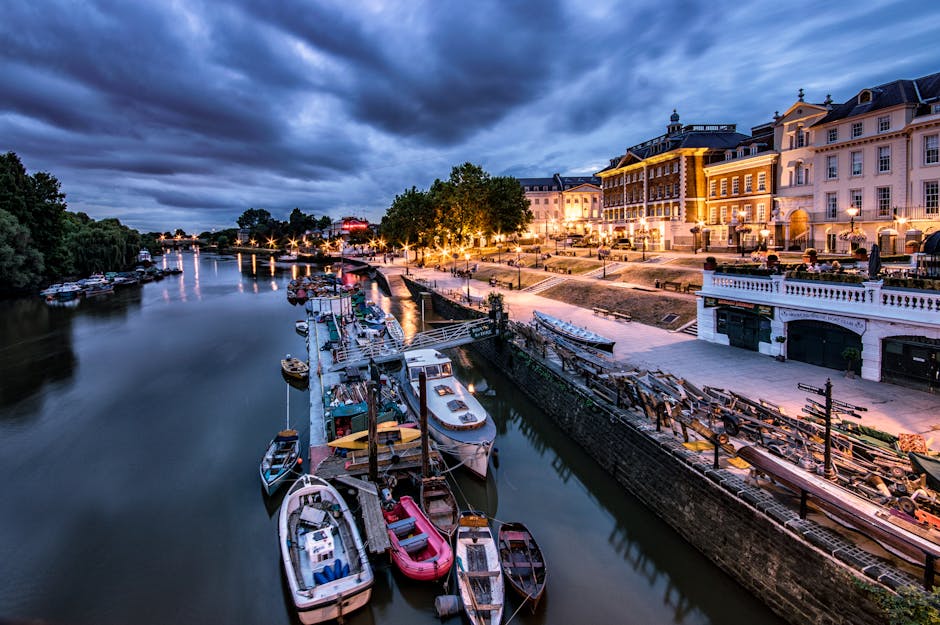A panoramic view of a riverside area in Kingston upon Thames during early evening, showing a calm waterway with several boats docked alongside the quay, some with protective covers. The boats vary in size and color, including white, blue, and red, and are moored close together. Adjacent to the water, there is a paved promenade with wooden benches, street lamps, and trees, all illuminated by warm lighting from nearby buildings. The background features multi-storey houses and shops with large windows, some of which are lit from within, showcasing a mix of historic and modern architecture. The site appears to be prepared for a home relocation or furniture transport operation, with some packing materials or equipment possibly visible on the pavement, illustrating the logistical aspect of house removals in Kingston upon Thames. The sky above is overcast with dark clouds, adding a dramatic atmosphere to the scene.