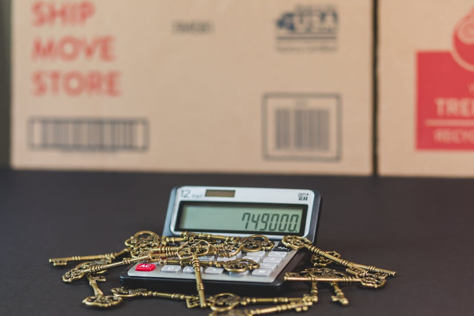 A digital calculator with a large display showing the number 749800 is placed on a black surface. Surrounding the calculator is a pile of gold-colored keys of various sizes and shapes, some with ornate bows and others with simpler designs. Behind the calculator and keys, there are three cardboard boxes stacked against a wall, with text such as 'SHIP MOVE STORE' and barcodes printed on them. The boxes appear to be used for packing belongings during a house relocation. The setting suggests the process of moving household items, involving packing, key management, and transportation. Occasionally, the scene reflects packing and moving logistics, with the presence of packing materials and items associated with furniture transport, relevant for a home removal service like those offered by removalvankingstonuponthames.co.uk.