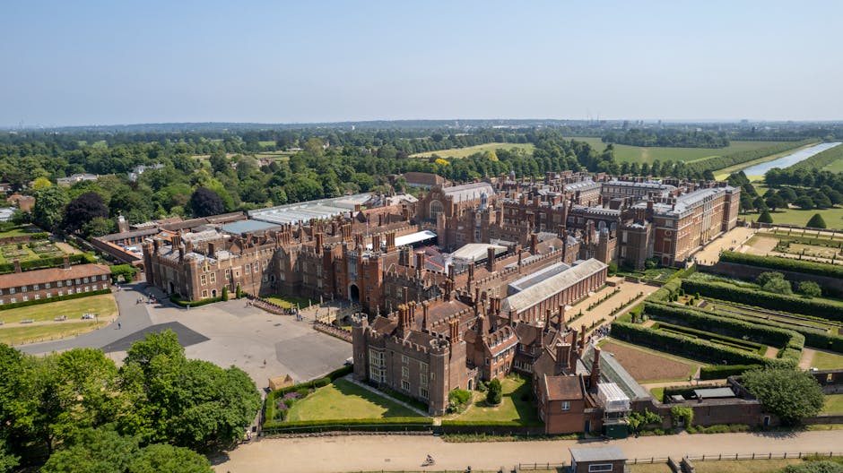 An aerial view of a large historic English estate with extensive brick buildings and ornate architectural details, surrounded by well-maintained gardens, lawns, and tree-lined pathways. In the foreground, a service van belonging to a removals company is parked on a paved driveway near an entrance gate, with several cardboard boxes and wrapped furniture pieces visible on the pavement and inside the vehicle. The van is in the process of being loaded or unloaded as movers, wearing casual clothing, carry belongings such as chairs, small cabinets, and wrapped household items across the driveway towards the estate's main entrance. The surrounding area features lush green fields and a river in the distance, under a clear sky with bright daylight. The scene captures a precise moment in the home relocation process, emphasizing careful packing and furniture transport for a house move in the Hampton Court area, aligning with the services offered by removalvankingstonuponthames.co.uk.