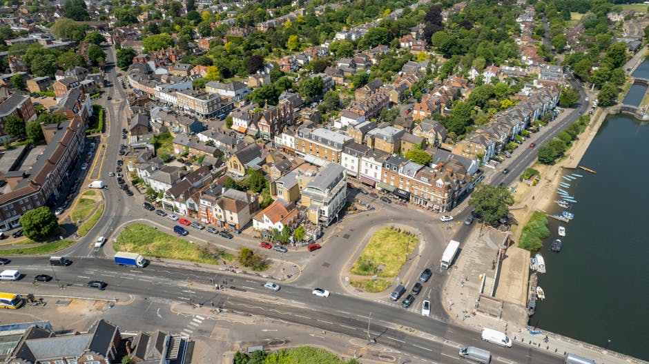 Aerial view of a coastal town with tightly packed residential buildings, including houses and small commercial structures, situated along a curving street and beside a body of water with small boats docked. The area features a mix of brick and clapboard facades, with some buildings having grey or white exteriors. The street shows moving traffic, including cars, trucks, and a bus, with a few parked vehicles along the road and near the waterfront. Adjacent to the buildings, there is a small, paved loading area with vehicles and a section of landscaped green space with grass and trees, possibly part of a home relocation or furniture transport process supported by [COMPANY_NAME]. The shoreline has a promenade or walkway, and the water is calm, reflecting the partly cloudy sky. The overall scene suggests a busy, scenic riverside or estuary town, suitable context for house removals, packing, and moving logistics in the Hampton Court area for Kingston homes.