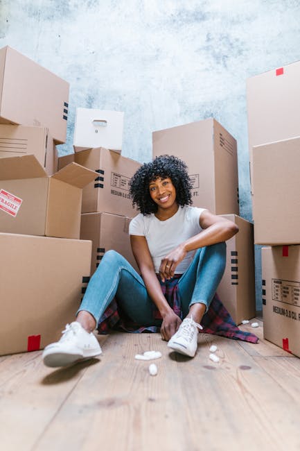 A young woman with curly black hair, smiling, sitting on the wooden floor of a room surrounded by numerous cardboard boxes, some sealed with packing tape and others partially open, indicating a home r