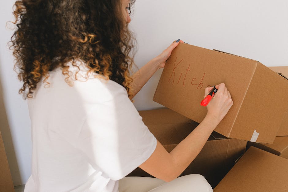 A woman with curly brown hair, wearing a white shirt, is inside a home near a doorway, preparing for a house move. She is writing the word 'Kitchen' in red marker on a medium-sized cardboard box, which is open and placed among other packed boxes. Several additional cardboard boxes are stacked around her, some sealed with packing tape, with labels or markings. The scene is well-lit with natural light, and the background features a plain white wall, indicating an indoor setting during packing and moving preparations. This image illustrates packing tasks associated with home relocation and the use of cardboard boxes for furniture transport and moving logistics, as part of professional removals services that may be offered by removalvankingstonuponthames.co.uk.