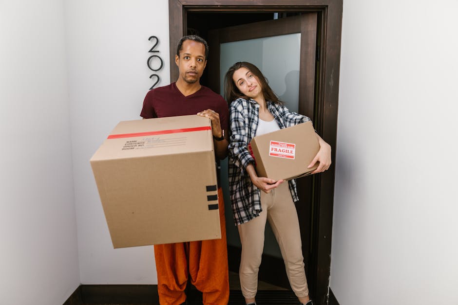 A woman with curly brown hair, wearing a white shirt, is inside a home near a doorway, preparing for a house move. She is writing the word 'Kitchen' in red marker on a medium-sized cardboard box, which is open and placed among other packed boxes. Several additional cardboard boxes are stacked around her, some sealed with packing tape, with labels or markings. The scene is well-lit with natural light, and the background features a plain white wall, indicating an indoor setting during packing and moving preparations. This image illustrates packing tasks associated with home relocation and the use of cardboard boxes for furniture transport and moving logistics, as part of professional removals services that may be offered by removalvankingstonuponthames.co.uk.