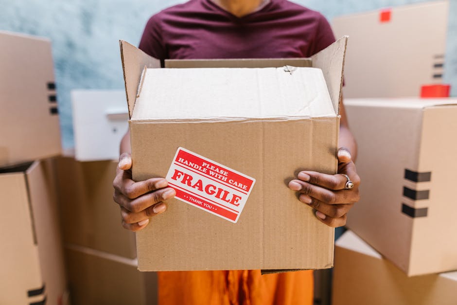 A person in a maroon shirt is holding an open cardboard box with both hands in an indoor setting, possibly a packing or storage area. The box is labeled with a red and white 'Fragile' sticker indicating careful handling, with the text 'Please handle with care' and 'Thank you' also visible. Surrounding the individual are several other cardboard boxes of varying sizes, some sealed and others open, indicating a packing or home relocation process. The environment appears well-lit with natural or fluorescent light, and the background includes a white door and additional boxes stacked or positioned on a flat surface, reflecting the typical scene of furniture transport, packing, or moving activities involving careful handling of items during a house removal or relocation process. This visual is associated with house removals and moving services, such as those provided by removalvankingstonuponthames.co.uk.