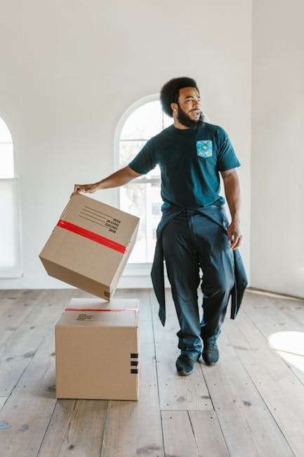 A male moving expert with dark curly hair and a beard is inside a room with white walls and a large window with an arched top, allowing natural daylight to illuminate the space. He is dressed in a navy blue t-shirt and dark trousers, with a relaxed posture. The man is holding a cardboard box secured with red packing tape in his right hand, preparing to load or unload it. On the wooden floor beside him, there is an additional cardboard box, also sealed with packing tape, indicating recent packing or unpacking activities as part of a home relocation. Behind him, a white wall and the window are visible, emphasizing the indoor environment. This scene captures a moment in the packing and moving process, typical of professional house removals, with the focus on handling and transporting packed boxes within a property, supporting efficient furniture transport and moving logistics. The image reflects careful attention to packing materials and tidy organization, which are key elements in successful residential removals, as seen in services provided by removalvankingstonuponthames.co.uk.