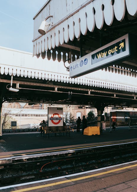 A view of Kingston Station platform in daylight, with a covered waiting area featuring a white canopy decorated with scalloped edges. The platform is occupied by several individuals, some standing and others sitting on benches, preparing for a home relocation. In the foreground, wooden furniture, cardboard boxes, and packing materials are visible, with some items being carried or positioned near the edge of a moving van. The van is parked adjacent to the platform, with its rear doors open, and yellow protective wheel stops are placed on the pavement. Above, a sign indicating the 'Way out' with an arrow points toward the station exit. The scene depicts a typical furniture transport and packing process in progress as part of a professional removals service by removalvankingstonuponthames.co.uk, supporting efficient moving logistics from the station area in Kingston upon Thames.