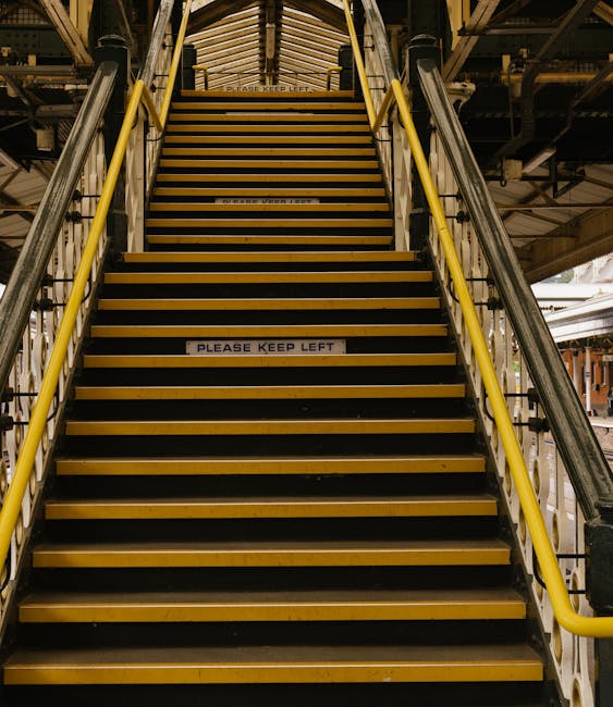 A set of industrial metal stairs with yellow handrails leading upward at a railway station, featuring black grip strips on each step and a sign reading 'PLEASE KEEP LEFT' attached to the lower part of the staircase. The surrounding environment includes metal scaffolding and overhead structures, indicating a location used for passenger access or freight movement. In the context of house removals or home relocation, this staircase could be part of a station used for transporting furniture or boxes during a moving service. The lighting is artificial, providing clear visibility of the steps and structural details, consistent with a busy station environment where careful navigation is essential for safe loading and unloading of packed household items.