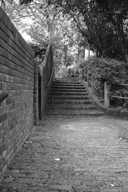 A narrow cobbled pathway leading uphill with a flight of uneven stone steps at the top, situated between a brick wall on the left and a railing on the right, surrounded by dense foliage and trees overhead. The stairs appear to have some parking or access issues due to the limited width and elevation, which could pose difficulties for large furniture or moving vans during a home relocation. In the foreground, the cobblestones are visible with scattered leaves and debris, indicating an outdoor environment. The scene may be part of a house removal or furniture transport process experienced on a tight street, typical of Kingston upon Thames, with natural lighting casting shadows through the trees, highlighting the textured surfaces of the steps and surrounding greenery. This setting aligns with logistical considerations for removals services provided by removalvankingstonuponthames.co.uk, especially in locations with restricted parking and narrow access points.