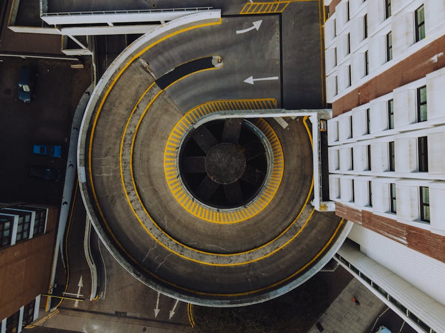 An aerial view showing a narrow, spiral underground car park ramp with concrete walls and yellow safety markings. The ramp is semi-circular, leading downward toward a central circular section that appears to be a structural support or ventilation feature. On the right side of the image, a white multi-storey building with numerous windows is visible, adjacent to the entrance of the ramp. The surrounding area includes a small section of pavement and parked vehicles on the upper left. This scene illustrates the logistical challenges of moving furniture or boxes through restricted access points, as often encountered during home relocation or furniture transport in areas like Kingston upon Thames, with the potential for parking issues and stairs highlighted in the context of removals services.