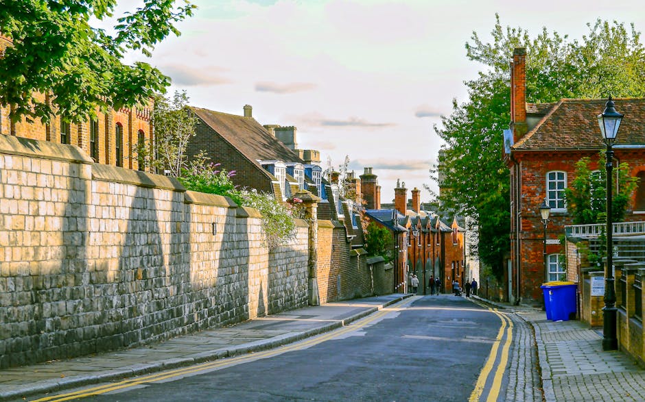 A narrow cobbled pathway leading uphill with a flight of uneven stone steps at the top, situated between a brick wall on the left and a railing on the right, surrounded by dense foliage and trees overhead. The stairs appear to have some parking or access issues due to the limited width and elevation, which could pose difficulties for large furniture or moving vans during a home relocation. In the foreground, the cobblestones are visible with scattered leaves and debris, indicating an outdoor environment. The scene may be part of a house removal or furniture transport process experienced on a tight street, typical of Kingston upon Thames, with natural lighting casting shadows through the trees, highlighting the textured surfaces of the steps and surrounding greenery. This setting aligns with logistical considerations for removals services provided by removalvankingstonuponthames.co.uk, especially in locations with restricted parking and narrow access points.