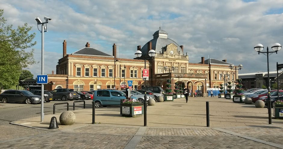 A large brick building with a Victorian architectural style, featuring arched windows, decorative stonework, and a central clock tower with a domed roof, situated above a busy parking area. In front of the building, there are cars parked along the pavement, with some being loaded or unloaded during a home relocation process. Several black bollards, decorative street lamps, and flower planters are aligned around the area, providing pedestrian guidance and aesthetic appeal. The pavement is made of light-colored concrete, and there are a few individuals walking, possibly involved in moving or packing items within nearby properties. The scene is well-lit with natural daylight, and the sky has patches of clouds, suggesting a clear, pleasant day. This setting is typical of the entrance to a railway station or commercial centre, relevant to the context of house removals and transport logistics, as managed by companies like removalvankingstonuponthames.co.uk for home relocation services.