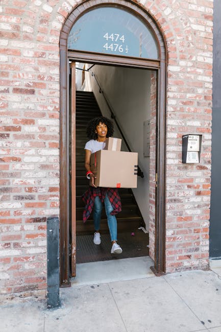 A woman with curly hair, wearing a white t-shirt, blue jeans, and white sneakers, is emerging from a brick property through a narrow arched doorway. She is carrying a large cardboard box with packing tape around its edges and a smaller box stacked on top. The doorway opens onto a staircase inside the building, with visible grey walls and wooden steps. To her right, an intercom system is mounted on the brick exterior wall. The scene is set during daytime, on a concrete sidewalk outside the property. This image depicts the process of home relocation and furniture transport, highlighting the careful handling and packing involved in professional removals, as performed by removalvankingstonuponthames.co.uk.