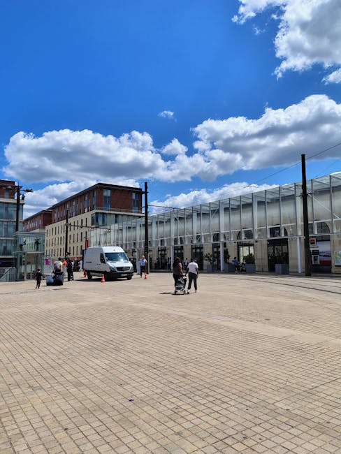 A large brick building with a Victorian architectural style, featuring arched windows, decorative stonework, and a central clock tower with a domed roof, situated above a busy parking area. In front of the building, there are cars parked along the pavement, with some being loaded or unloaded during a home relocation process. Several black bollards, decorative street lamps, and flower planters are aligned around the area, providing pedestrian guidance and aesthetic appeal. The pavement is made of light-colored concrete, and there are a few individuals walking, possibly involved in moving or packing items within nearby properties. The scene is well-lit with natural daylight, and the sky has patches of clouds, suggesting a clear, pleasant day. This setting is typical of the entrance to a railway station or commercial centre, relevant to the context of house removals and transport logistics, as managed by companies like removalvankingstonuponthames.co.uk for home relocation services.