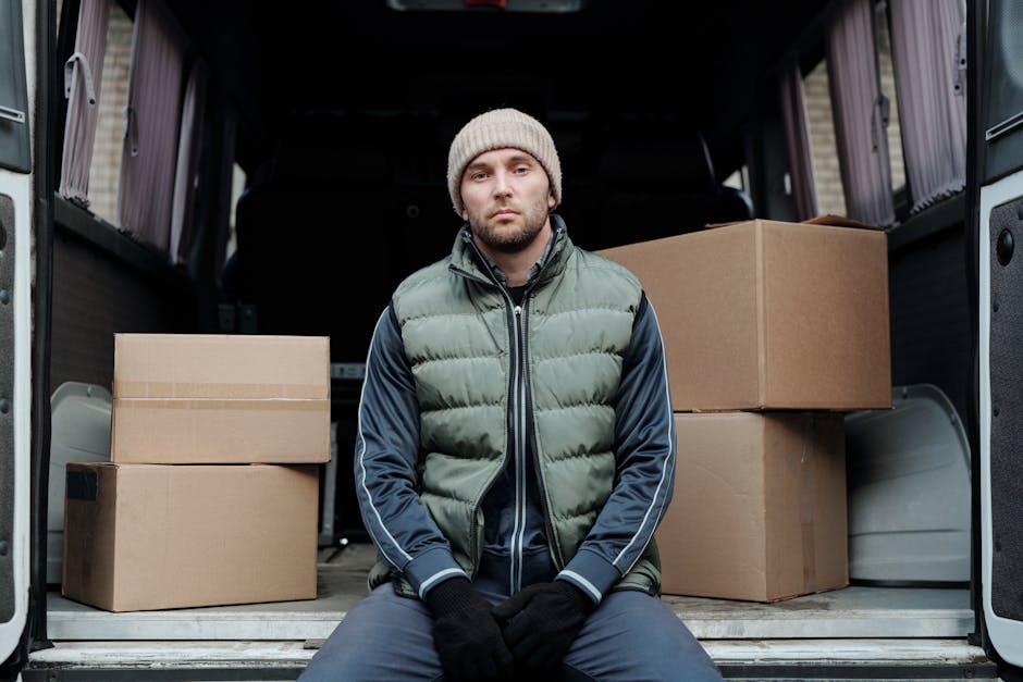 A young man wearing a beige knit hat, a green and navy padded jacket, and black gloves is sitting on the edge of an open moving van. Behind him, the van's interior reveals several packed cardboard boxes, some sealed with tape and others stacked on top of each other, indicating a home relocation process. The boxes vary in size and are placed on the vehicle’s floor, with larger boxes positioned toward the back and smaller ones in front. The van’s interior walls are visible, showing a dark finish, and the rear doors are open, exposing a paved area outside. The natural lighting highlights the details of the boxes, the man's clothing, and the surrounding environment. This scene is part of a furniture transport or packing and moving operation, possibly managed by [COMPANY_NAME], which offers removals services in Kingston upon Thames. The context reflects the loading or unloading stage of a house removal, emphasizing the logistical aspects of a professional move for a residential relocation.